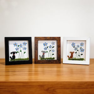 May include: Three framed artworks depicting dog silhouettes amongst blue flower illustrations. The frames are black, brown, and white, each with a different coloured dog. The artwork is set against a white background with green moss.