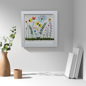 May include: A framed floral artwork featuring colorful flowers, a butterfly, and a bee. The artwork is in a white frame and is displayed on a white wall. A vase with greenery and a small cork container are in the foreground.