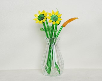 May include: A clear glass vase holds a bouquet of glass flowers. Two yellow sunflowers with green centers and stems are the focal point. An orange, spiky flower and green leaves complete the arrangement. The vase has a geometric design.