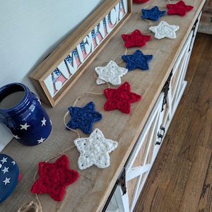 May include: A wooden sign with the word "AMERICA" in red, white, and blue. A garland of red, white, and blue crocheted stars is draped across a wooden surface. A blue jar with white stars and a red and white striped jar are also visible.