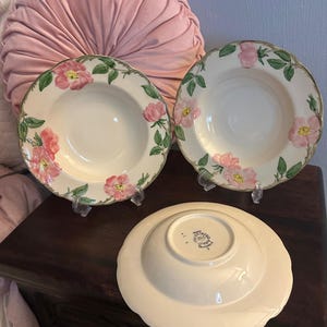 May include: A white ceramic mug with a black handle and the text "But First, Coffee" in a black, bold font. The mug is on a white surface. The mug is approximately 10 cm tall.