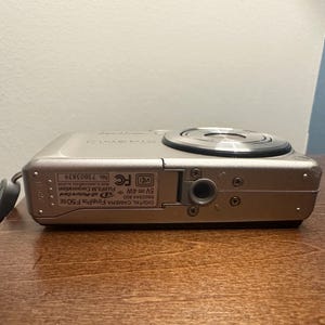 May include: A silver Fujifilm FinePix F50SE digital camera. The camera has a lens on the top and a small strap attached to the side. The camera is resting on a wooden surface.