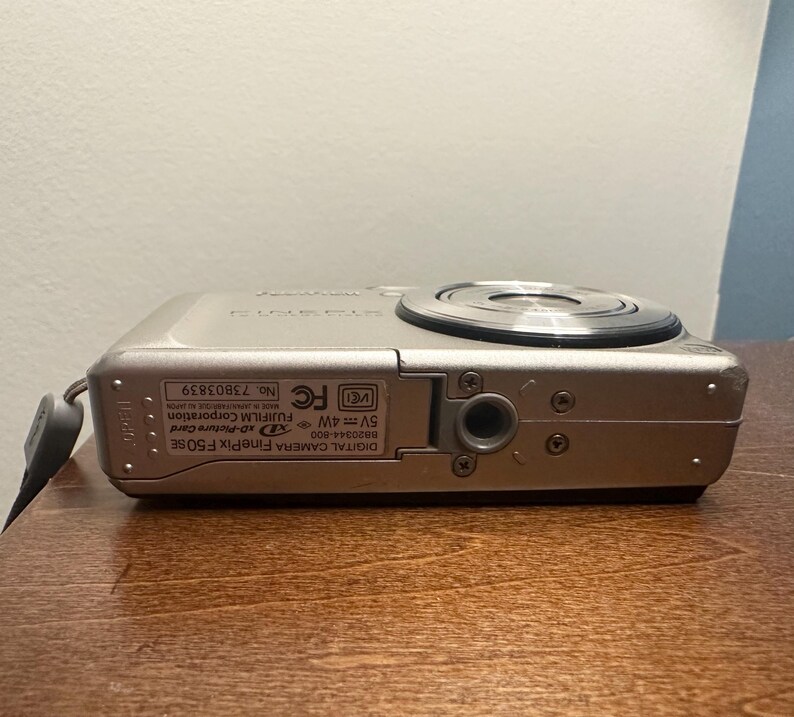 May include: A silver Fujifilm FinePix F50SE digital camera. The camera has a lens on the top and a small strap attached to the side. The camera is resting on a wooden surface.