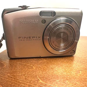 May include: A silver Fujifilm FinePix digital camera. The camera has a zoom lens and a built-in flash. The text "FUJIFILM", "FINEPIX", and "12.0 MEGA PIXELS" are visible on the camera body. The lens reads "FUJINON ZOOM LENS 3x f3.8-24mm 1:2.8-5.1".