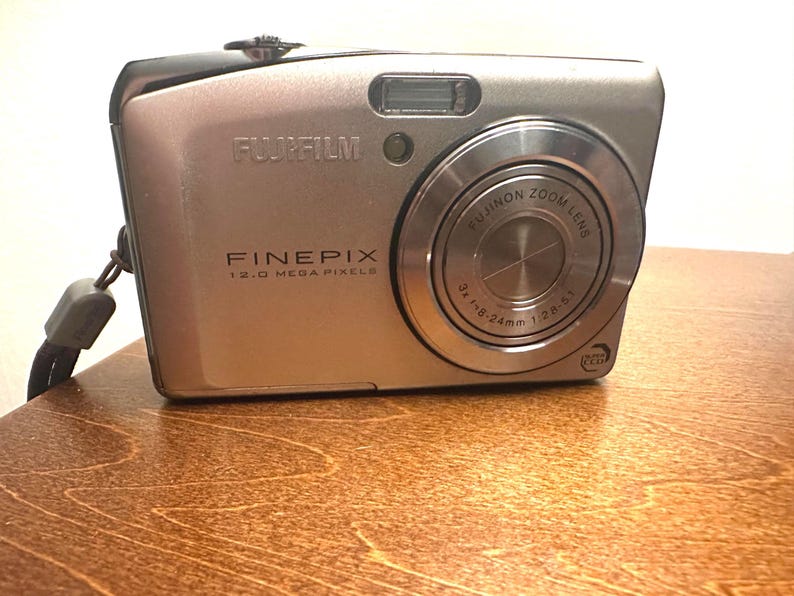 May include: A silver Fujifilm FinePix digital camera. The camera has a zoom lens and a built-in flash. The text "FUJIFILM", "FINEPIX", and "12.0 MEGA PIXELS" are visible on the camera body. The lens reads "FUJINON ZOOM LENS 3x f3.8-24mm 1:2.8-5.1".