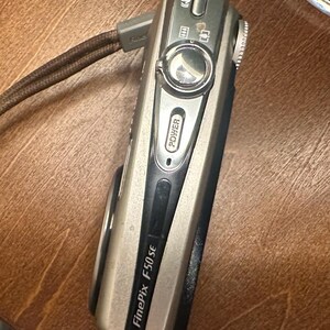 May include: A silver and black FinePix F50 SE digital camera with a brown wrist strap. The camera has a power button and several other function buttons. The camera is on a wooden surface.