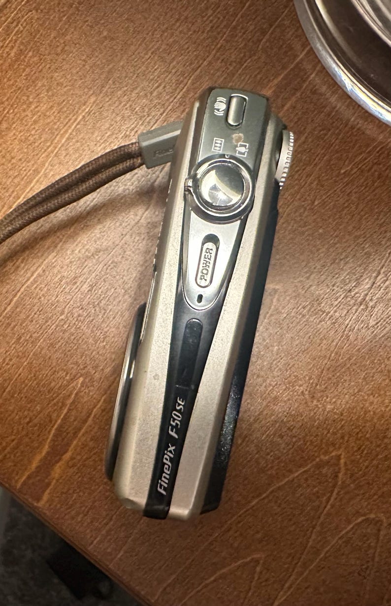 May include: A silver and black FinePix F50 SE digital camera with a brown wrist strap. The camera has a power button and several other function buttons. The camera is on a wooden surface.