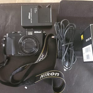 May include: A black Nikon digital camera with a lens, strap, charger, and battery. The camera strap is black with yellow text that reads "Nikon". The battery charger is labeled "MH-24". The battery box is labeled "EN-EL14".