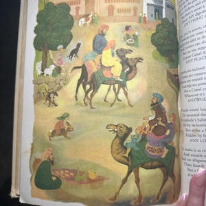 May include: An illustrated page from a children's book depicts a desert scene with camels carrying people. The artwork features a detailed illustration of a town with buildings and people in traditional clothing. The text on the right side of the page is partially visible.