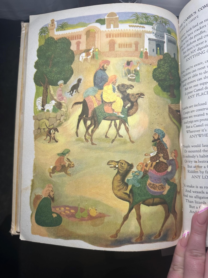 May include: An illustrated page from a children's book depicts a desert scene with camels carrying people. The artwork features a detailed illustration of a town with buildings and people in traditional clothing. The text on the right side of the page is partially visible.