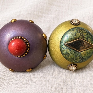 May include: Two spherical drawer knobs. The left knob is matte purple with small gold studs and a central red bead. The right knob is metallic gold with a green textured circular inlay, a central black diamond shape, and small clear crystal accents.