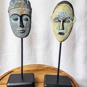 May include: Two sculpted face masks mounted on thin black metal rods and square black bases. The left mask is blue-grey with a textured surface and a triangular patterned headband. The right mask is yellow with a dark forehead and textured finish.