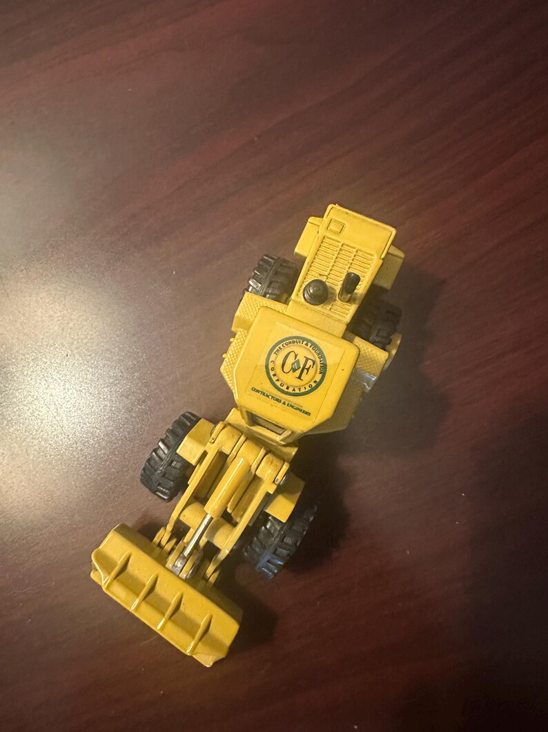 May include: A yellow toy construction vehicle featuring a front loader bucket, black rubber tyres, and a circular logo on the top hood that reads C&F Corporation, The Conduit & Foundation Corporation, Contractors & Engineers in black text.