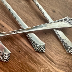 May include: Three silver-coloured metal spoons featuring a floral rose motif at the handle ends. The handles are engraved with the text Damask Rose and Heirloom Sterling. The spoons have a polished finish and a tapered design with detailed rose carvings.