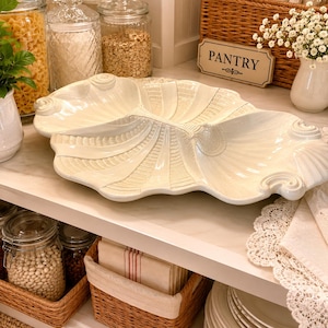 May include: A cream-coloured ceramic serving dish shaped like a large, elongated seashell with three distinct sections. The surface features intricate, raised radial ridges and decorative scrollwork at the ends, finished with a glossy glaze.