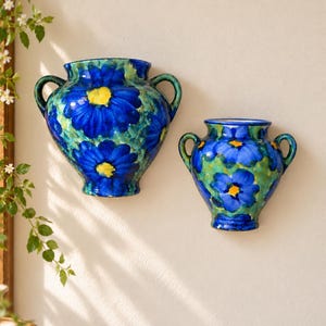 May include: Two ceramic wall-mounted vases featuring a hand-painted floral pattern with vibrant blue petals, yellow centers, and green foliage accents. Each vessel has a rounded body, a wide opening, and two curved side handles.