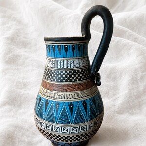 May include: A ceramic pitcher featuring a tall black handle and a rounded body decorated with horizontal bands of geometric patterns in shades of bright blue, black, white, and brown. The design includes triangles, checkerboards, and lines.