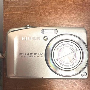 May include: Silver Fujifilm FinePix digital camera with a Fujinon zoom lens. The camera has a 12.0 megapixel resolution and a 3x zoom lens. The camera is compact and has a built-in flash.