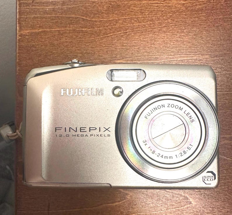 May include: Silver Fujifilm FinePix digital camera with a Fujinon zoom lens. The camera has a 12.0 megapixel resolution and a 3x zoom lens. The camera is compact and has a built-in flash.