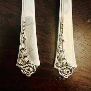 Op de afbeelding: Close-up van twee zilverkleurige gebruiksvoorwerpen. Elk gebruiksvoorwerp heeft een bloemenroosontwerp aan de basis en decoratieve bladeraccenten. De gebruiksvoorwerpen liggen op een donkerbruin houten oppervlak.