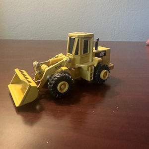 May include: A yellow Caterpillar 950E wheel loader toy vehicle featuring a front bucket, black tyres, and a cabin with dark windows. The side of the loader arm is marked with the text 950E, and the rear engine housing displays the CAT logo.
