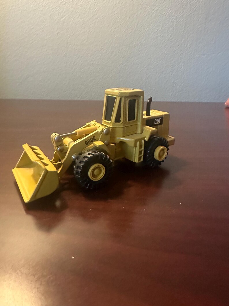 May include: A yellow Caterpillar 950E wheel loader toy vehicle featuring a front bucket, black tyres, and a cabin with dark windows. The side of the loader arm is marked with the text 950E, and the rear engine housing displays the CAT logo.