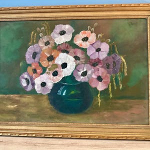 May include: An oil painting in a gold frame depicting a bouquet of anemone flowers in shades of white, pink, and purple. The flowers are arranged in a dark green rounded vase set against a muted green background on a tan surface.