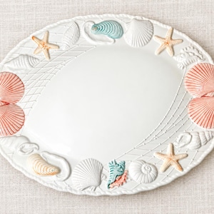 May include: An oval white ceramic serving platter featuring a raised relief border of various seashells, including pink scallops and orange starfish, integrated with a delicate net pattern that frames the smooth, plain center of the dish.