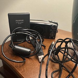 May include: A black Fujifilm BC-50 battery charger and a digital camera with black cables. The charger is labeled "FUJIFILM BATTERY CHARGER BC-50". The camera is black and silver.