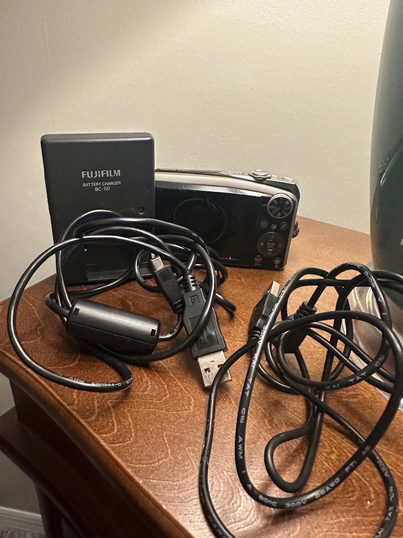 May include: A black Fujifilm BC-50 battery charger and a digital camera with black cables. The charger is labeled "FUJIFILM BATTERY CHARGER BC-50". The camera is black and silver.
