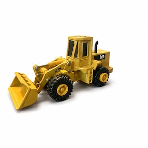 May include: A yellow CAT 950E wheel loader toy vehicle featuring a front bucket, black tyres with yellow hubs, and a cabin with black windows. The construction equipment model displays the CAT logo on the side and the model number 950E on the arm.
