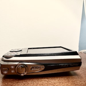 May include: A silver and black Fujifilm FinePix F50fd digital camera. The camera is resting on a wooden surface. The camera has a lens, buttons, and a small screen. The camera is a compact point-and-shoot model.