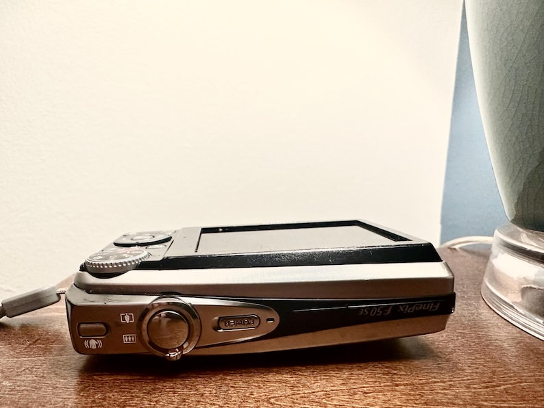 May include: A silver and black Fujifilm FinePix F50fd digital camera. The camera is resting on a wooden surface. The camera has a lens, buttons, and a small screen. The camera is a compact point-and-shoot model.