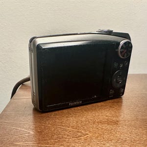 May include: A black Fujifilm digital camera with a silver trim, resting on a wooden surface. The camera's screen is black, and the Fujifilm logo is visible. The camera has a dial and several buttons.