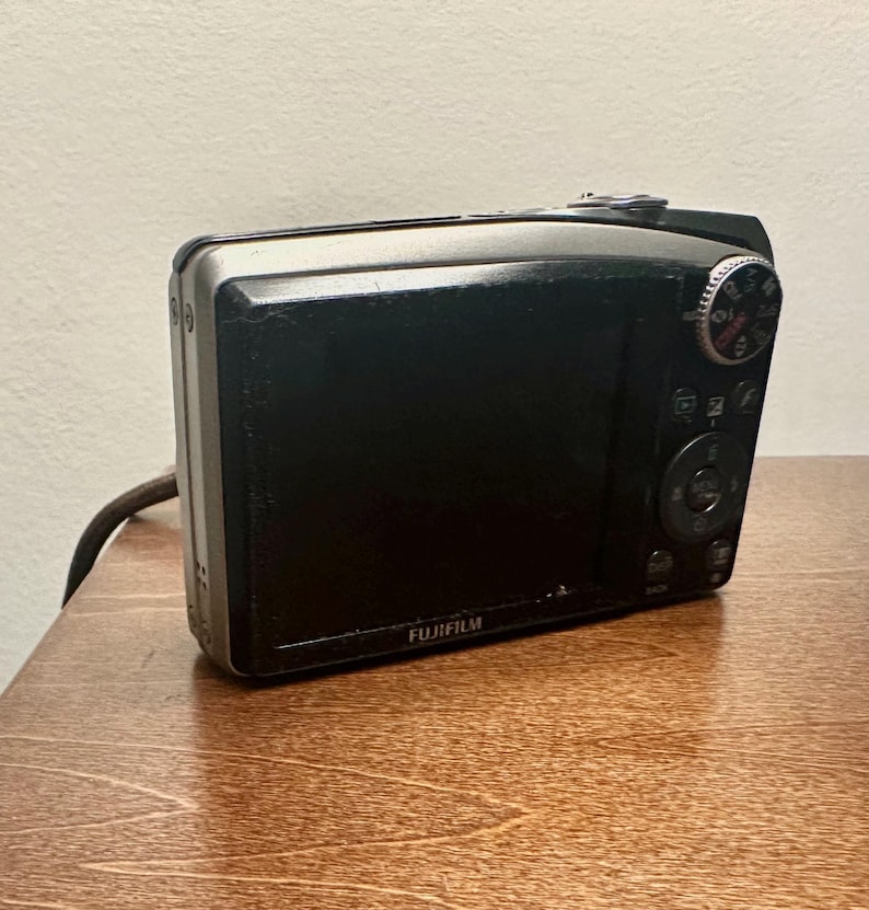 May include: A black Fujifilm digital camera with a silver trim, resting on a wooden surface. The camera's screen is black, and the Fujifilm logo is visible. The camera has a dial and several buttons.