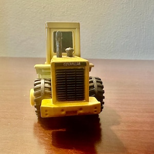 May include: A yellow Caterpillar toy tractor featuring a black front grille, a vertical exhaust pipe, and black rubber tyres. The front of the vehicle displays the brand name Caterpillar in black lettering on a rectangular label above the grille.