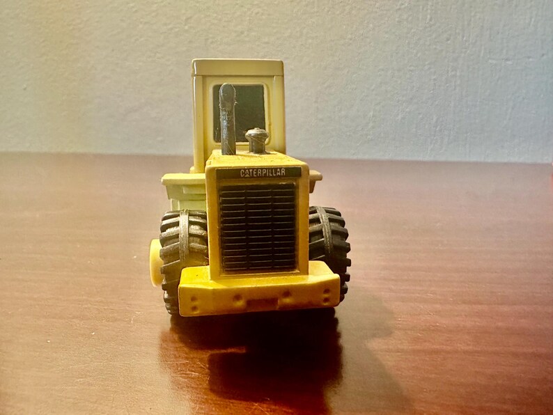 May include: A yellow Caterpillar toy tractor featuring a black front grille, a vertical exhaust pipe, and black rubber tyres. The front of the vehicle displays the brand name Caterpillar in black lettering on a rectangular label above the grille.