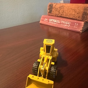 May include: A yellow die-cast toy wheel loader featuring a front bucket, a central operator cab with black windows, and four black treaded tyres. The top of the cab displays a circular logo with the letters CF printed in the centre.