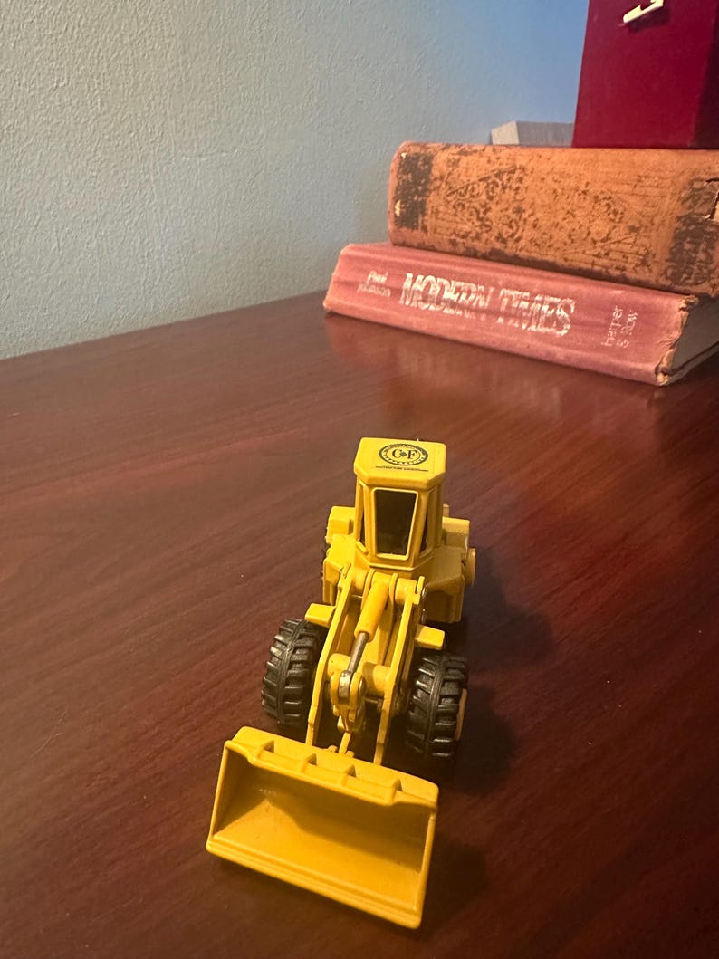 May include: A yellow die-cast toy wheel loader featuring a front bucket, a central operator cab with black windows, and four black treaded tyres. The top of the cab displays a circular logo with the letters CF printed in the centre.