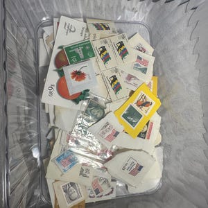 Könnte beinhalten: Eine transparente Kunststoffbox gefüllt mit einer Sammlung farbenfroher Briefmarken. Die Briefmarken zeigen verschiedene Designs, darunter Flaggen, Blumen und Porträts. Einige Briefmarken tragen Text, darunter "USA 23". Die Box hat einen goldenen Griff.