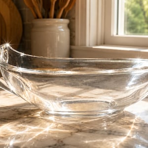 May include: A clear glass gravy boat with a smooth, curved silhouette, a single handle on one side, and a tapered pouring spout on the opposite end. The vessel features a wide, open top and a rounded base, reflecting light across its transparent surface.