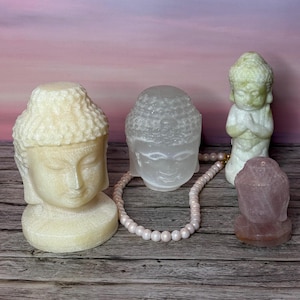 May include: A collection of Buddha statues in various colors and sizes. The statues include a cream-colored Buddha head, a clear Buddha head, a green Buddha statue with hands together, and a pink Buddha head. A pearl necklace is draped between the statues.