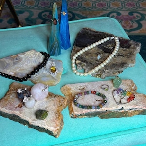 May include: An assortment of gemstone jewelry, including necklaces, bracelets, and rings, displayed on natural stone slabs. The jewelry features various colors and textures, with a focus on pearls, beads, and pendants. The background is a light blue surface.