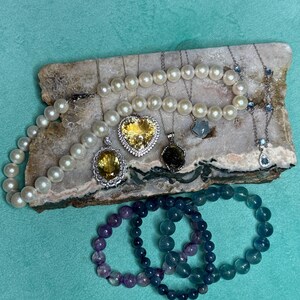 May include: An assortment of jewelry including pearl necklaces, gemstone pendants, and beaded bracelets. The pendants feature yellow and green gemstones, and the bracelets are in shades of purple and blue. The jewelry is displayed on a stone slab.