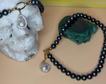 Può includere: Due collane con perle grigio scuro. Una collana ha una chiusura dorata e un ciondolo a forma di goccia di perla con un bordo simile a un diamante. L'altra collana è parzialmente visibile. Le collane sono esposte su un teschio bianco e una pietra verde.