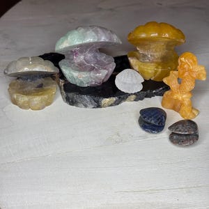 Puede incluir: Una colección de esculturas de piedras preciosas talladas, que incluyen formas de hongo en tonos amarillos, verdes y morados. También, una pequeña figura naranja y varias conchas de piedra pulida pequeñas se exhiben sobre una superficie oscura.