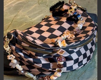 Puede incluir: Un bolso de cintura a cuadros negros y beige con cremallera y herrajes dorados. La bolsa está adornada con una correa de cuentas con abalorios de setas, mariposas y flores. Un abalorio negro y esponjoso y otros abalorios decorativos están adjuntos.