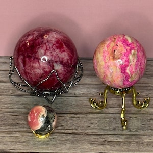 May include: Three gemstone spheres of varying sizes and colors. The largest is a deep red sphere in a black metal stand. The second is a pink and cream sphere on a gold stand. The smallest is pink, black, and cream on a gold stand.