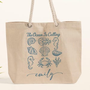 May include: Beige canvas tote bag with rope handles. The bag features blue illustrations of seashells and marine life, with the text "The Ocean Is Calling" and a name in cursive. A palm leaf is in the background.
