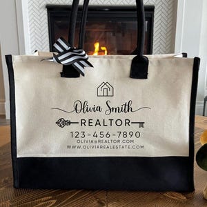 Peut inclure: Un sac fourre-tout en toile avec des poignées noires et un fond noir. Le sac affiche le texte "Olivia Smith, Realtor" avec une maison et une clé. Un nœud rayé noir et blanc est attaché à la poignée.
